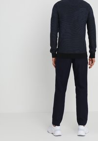 Marinblå tröja med texturerat mönster och ribbade muddar, kombinerad med mörk marinblå byxor. Modellen står med ryggen mot betraktaren. Vita sneakers syns.