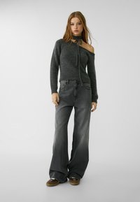 Dunkelgrauer Off-Shoulder-Pullover mit Bindungen am Hals, kombiniert mit hellgrauen weiten Jeans und braunen Sneakers. Weiche Textur, lässige Passform.