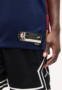 Κοντινό πλάνο ενός ατόμου που φοράει μπλε σκούρα φανέλα NBA και μαύρα σορτς μπάσκετ με λευκές ρίγες, δείχνοντας την αυθεντική ετικέτα Nike της φανέλας.