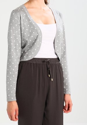 Femme portant un cardigan court gris à pois blancs sur un débardeur blanc et un pantalon noir ample avec cordon de serrage.