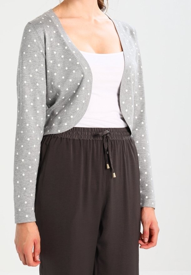 Femme portant un cardigan court gris à pois blancs sur un débardeur blanc et un pantalon noir ample avec cordon de serrage.