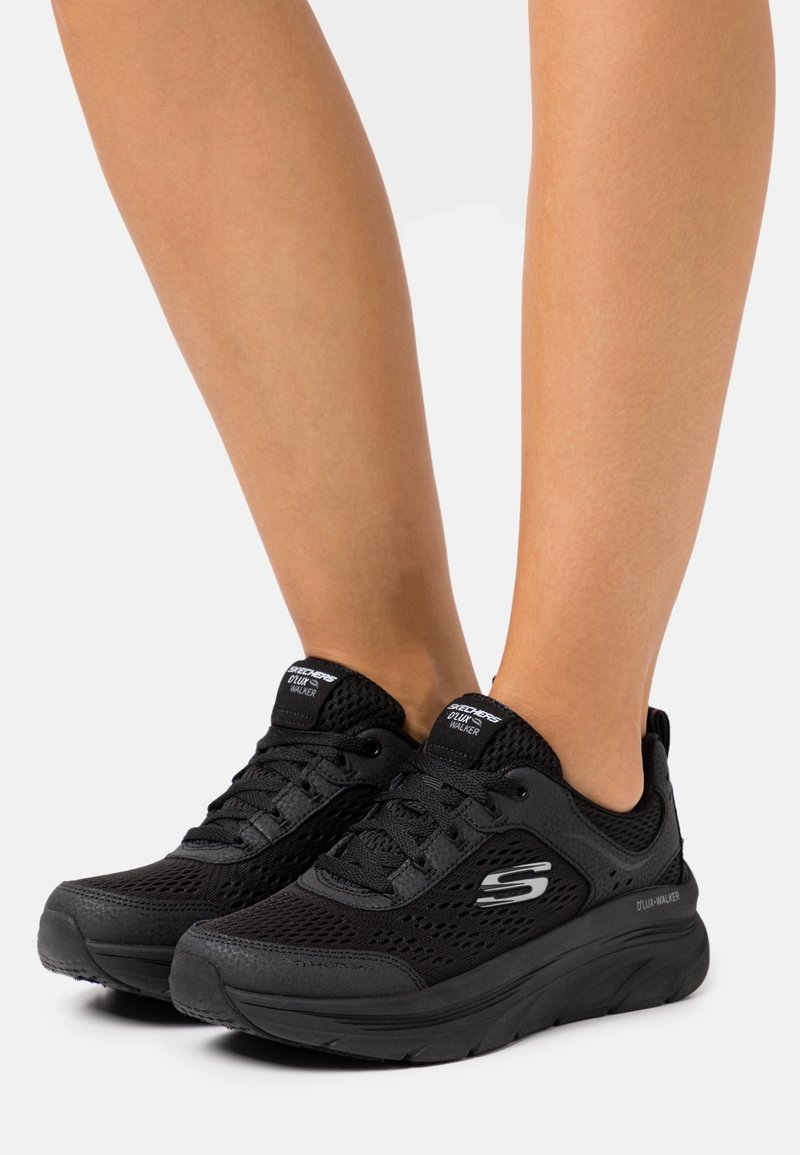 Jambes portant des baskets en mesh noires Skechers D'LUX Walker avec des semelles épaisses, debout sur un fond clair uni.