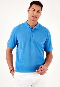 Camisa polo azul con un cuello clásico, tapeta de dos botones, mangas cortas y una tela texturizada. Llevada con pantalones cortos de color claro.