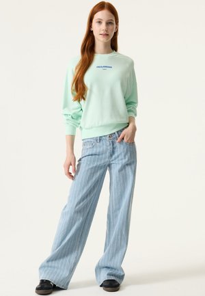 Jonge vrouw met lang rood haar die een muntgroene trui en lichtblauwe jeans met wijde pijpen en strepen draagt, staand met één hand in haar zak.