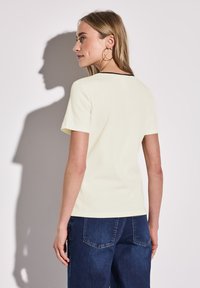 T-shirt a maniche corte a coste di colore giallo pallido, con colletto nero. Modello che indossa jeans in denim blu, in piedi con la schiena rivolta.