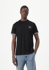 T-shirt de algodão preto com gola redonda, mangas curtas com riscas brancas e um pequeno patch retangular com logótipo no peito.