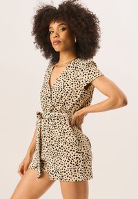Short-sleeve romper in beige with black leopard print. Features a deep V-neck and a belted waist for a defined silhouette. Soft fabric texture.