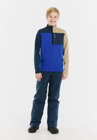 Giacca in pile blu e beige con zip e maniche lunghe, abbinata a pantaloni impermeabili blu scuro e stivali neri, indossata da un bambino sorridente.