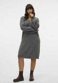 Vestido de suéter gris de punto con escote en V, mangas largas acanaladas y corte holgado, combinado con botas marrones hasta el tobillo. Textura suave, hasta la rodilla.