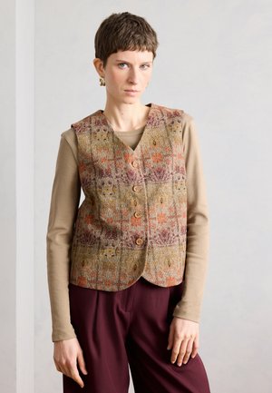 Žena s krátkymi vlasmi, ktorá má zlaté náušnice, vzorovanú zapínaciu vestu, béžový dlhý rukáv a bordové nohavice, stojaca pri obyčajnej stene.