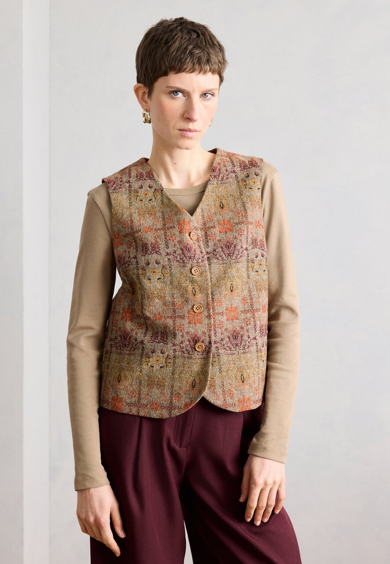 Γυναίκα με κοντά μαλλιά που φορά χρυσά σκουλαρίκια, ένα εμπριμέ γιλέκο με κουμπιά, μπεζ μπλούζα με μακριά μανίκια και μπορντό παντελόνια, στέκεται μπροστά σε έναν απλό τοίχο.