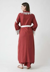Robe longue de couleur rouille en tissu texturé avec des franges aux manches et au col, cintrée à la taille par une ceinture blanche. Design doux et fluide.