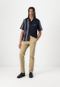 Camisa de manga corta en color azul marino con patrones de rayas horizontales y un logo en el frente. Combinada con pantalones caqui claros y mocasines negros.