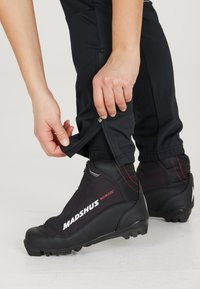 Mustat pohjoismaiseen hiihtoon tarkoitetut saappaat punaisilla yksityiskohdilla, teksturoitu päällinen ja ergonominen muotoilu. Henkilö säätää vetoketjua housujen helmasta saappaiden vieressä.