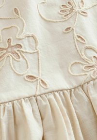 Hellbeiges Stoffmaterial mit filigraner Blütenstickerei in Hellbraun, strukturierter Oberfläche und gerafftem Rüschensaum am Saum.