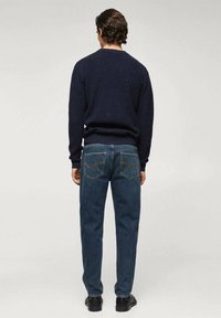 Mężczyzna stojący tyłem, ubrany w ciemnoniebieski sweter, niebieskie dżinsy i czarne buty, na jednolitym jasnym tle.