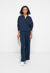 Marineblauwe sweatshirt met een kraag en een ontspannen pasvorm, gecombineerd met marineblauwe wijde broek met witte zijstrepen. Witte schoenen in de hand.