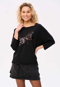 Zwarte oversized top met een pailletten libelleontwerp, lange mouwen en een brede ronde halslijn. Gecombineerd met een donkere denim rok met zakken.