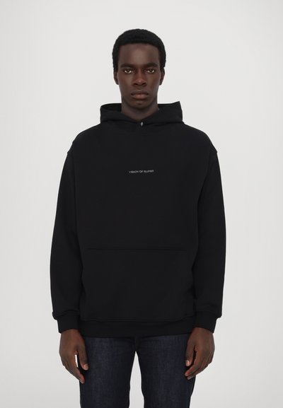 Sudadera negra de tejido suave con un bolsillo tipo canguro, que presenta un pequeño logo de texto blanco "VISION OF SUPER" en el pecho. Jeans de mezclilla oscura.