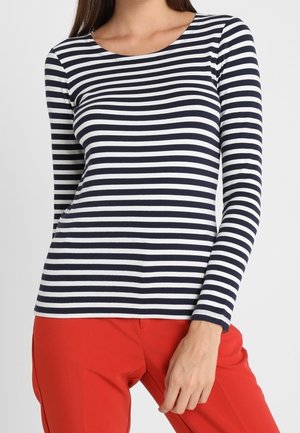 Femme portant un t-shirt à manches longues rayé horizontalement en noir et blanc et un pantalon rouge vif, la main reposant sur sa cuisse.