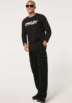 Czarna koszula z długim rękawem z logo "OAKLEY", w połączeniu z czarnymi spodniami cargo z kieszeniami i czarnymi sneakersami. Model nosi okulary przeciwsłoneczne.