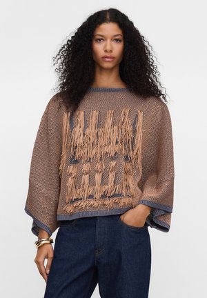 Frau mit lockigem Haar trägt einen lockeren braunen Strickpullover mit Fransen und dunkelblaue Jeans, posiert mit einer Hand in der Tasche.