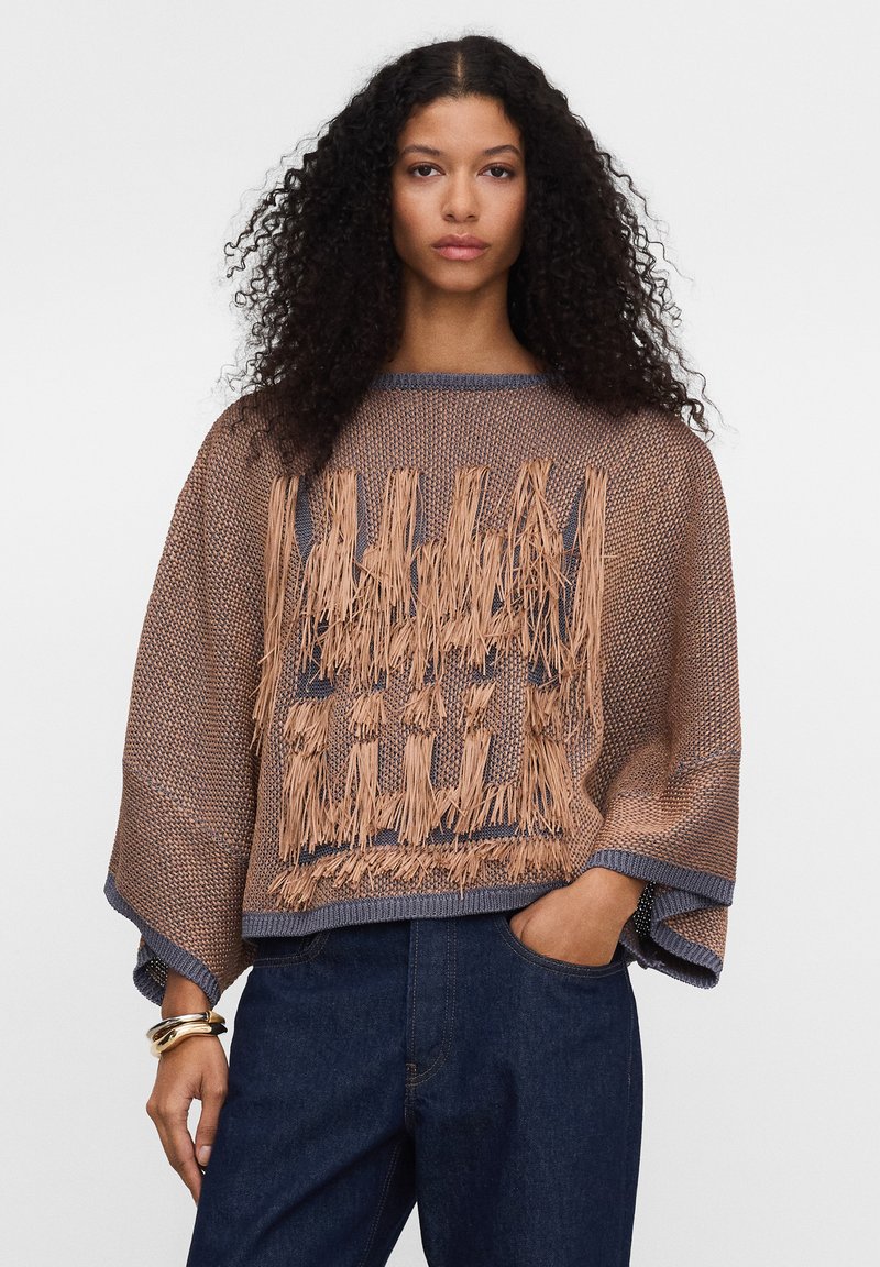 Kobieta o kręconych włosach w luźnym brązowym swetrze z frędzlami i ciemnoniebieskich dżinsach, pozuje z jedną ręką w kieszeni.