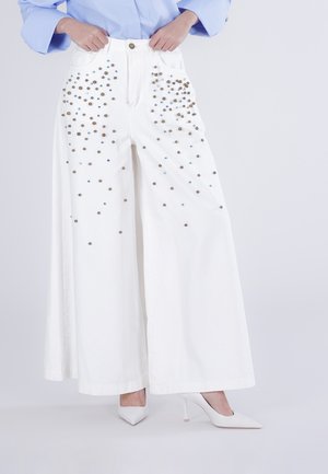 Pantaloni bianchi a gamba larga con borchie dorate e blu sparse, abbinati a décolleté bianche a punta e una camicetta azzurro chiaro.