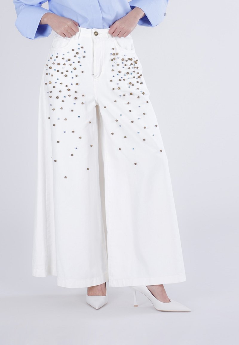 Hvoremte, hvide bukser med brede ben og spredte guld- og blå nitter, stylet med hvide, spidse højhælede sko og en lyseblå bluse.