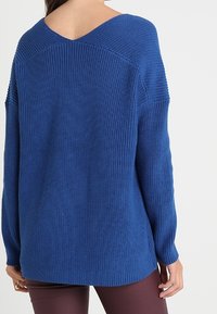 Maglione blu a maglia con ampia scollatura a V, tessuto testurizzato, spalle cadute e maniche lunghe, con orlo posteriore leggermente più lungo.