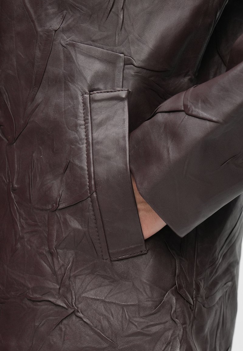 Manteau en cuir bordeaux avec une texture froissée, comportant une poche latérale et une finition douce et lisse. Manches longues avec poignets droits.