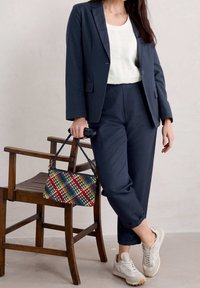 Navy tailored blazer and matching pants, paired with a white top and beige sneakers, holding a multicolored woven handbag. Wooden chair in background.