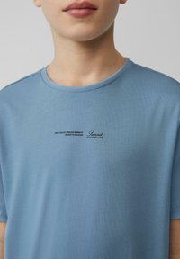 Lichtblauw katoenen t-shirt met een geribbelde ronde hals. Voorzien van zwarte bedrukte tekst op de voorkant. Korte mouwen, gladde textuur.