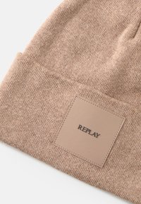 Strikket beanie i lysebrunt med et tekstureret mønster. Har et firkantet lædermærke prægnet med "REPLAY" i sort.