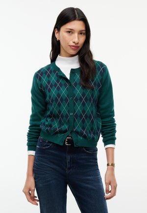 Kvinde iført en grøn argyle knapcardigan over en hvid rullekrave og mørkeblå jeans, stående mod en ensfarvet hvid baggrund.