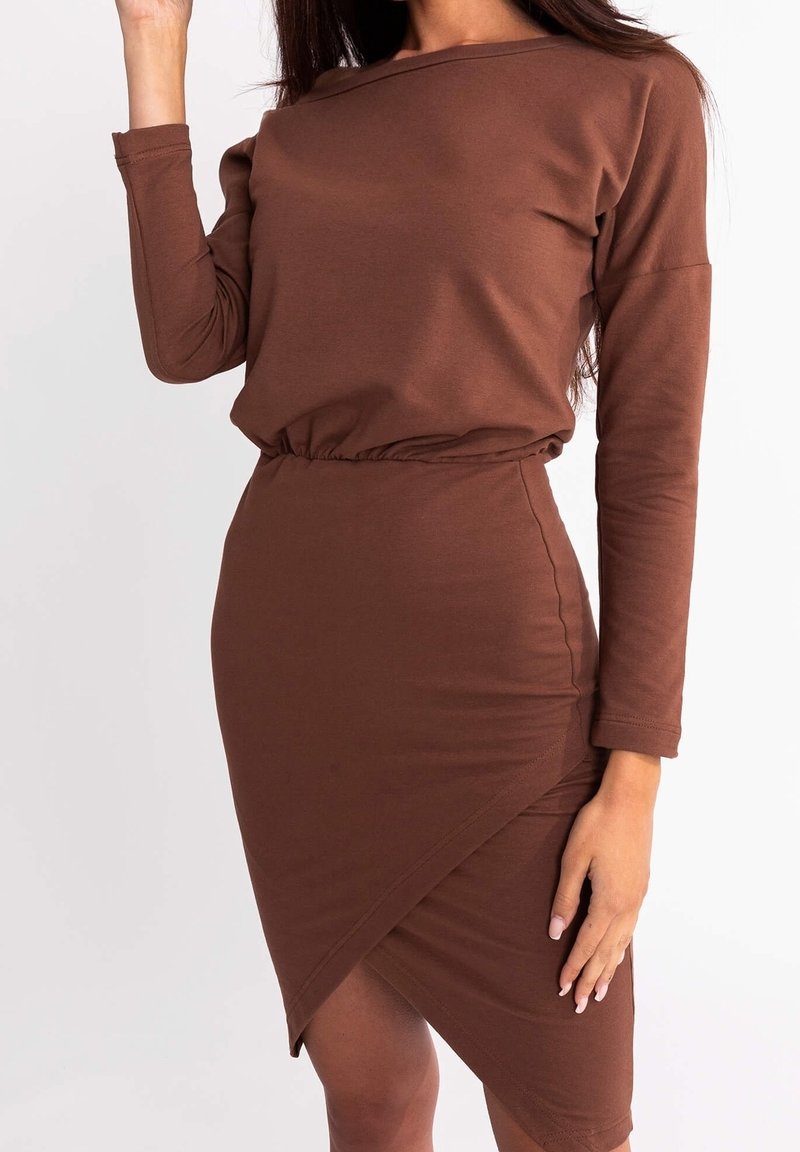 Femme portant une robe longue marron ajustée avec une taille froncée et un ourlet asymétrique sur un fond blanc uni.