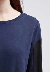 Woman wearing a navy blue sweater with a round neckline and black fabric detail on the sleeve.