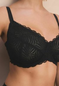 Svart spets-bh med blomstermönster, vågiga kanter och justerbara remmar. Designad med en mjuk textur och underwire-stöd.