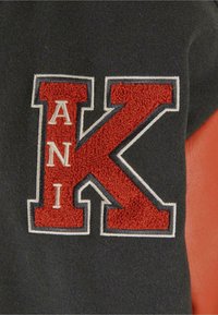 Svart stoff med en fremtredende rød og hvit brodert "K" og teksten "ANI," med et teksturert løkkemønster. Glatt oransje skinn til høyre.
