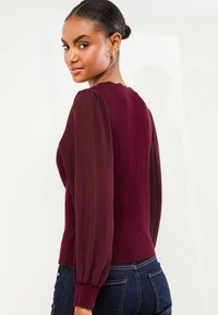 Bordeaux-farbenes, geripptes Sweater mit durchsichtigen Ballonärmeln, rundem Halsausschnitt und tailliertem Schnitt. Glatte Textur mit einem kontrastierenden Stoffdetail an den Ärmeln.