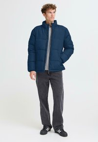 Marineblå puffjakke med høy krage, horisontal quiltet design og sidelommer, båret over en grå skjorte og kombinert med mørke jeans.