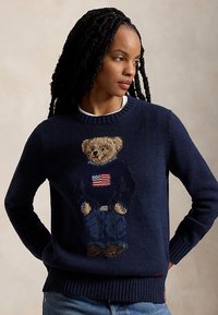 Vrouw met lange vlechten draagt een marineblauwe trui met een teddybeer gekleed in spijkerbroek en een trui met een ontwerp van de Amerikaanse vlag.