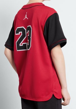 Personne portant un maillot de sport rouge et noir avec le numéro 23 et un logo Jumpman blanc dans le dos, debout devant un fond uni.