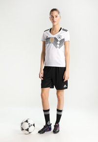 Witte voetbalshirt met zwarte horizontale strepen en embleem, gecombineerd met zwarte shorts en kniehoge zwarte sokken. Een voetbal ligt tussen haar voeten.