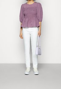 Femme portant un chemisier froncé violet, un pantalon blanc, des baskets blanches, tenant un petit sac à main lavande avec une courte lanière.