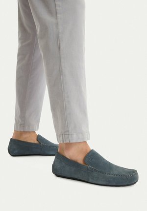 Persona che indossa pantaloni grigio chiaro e mocassini in camoscio blu, in piedi su uno sfondo chiaro, piedi mostrati da angoli laterali e posteriori.