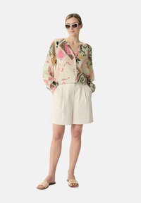Bluzka w kwiatowy wzór w pastelowych kolorach, zestawiona z beżowymi, plisowanymi szortami oraz plecionymi sandałami na płaskiej podeszwie, uzupełniona dużymi okularami przeciwsłonecznymi.