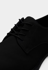 Chaussure habillée en cuir noir avec bout rond, laçage fermé et finition mate lisse sur fond blanc.