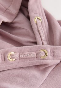 Weicher mauvefarbener Strickstoff mit passenden Kordeln, die durch goldene Ösen an einem Kleidungsstück gezogen sind, mit dezent gedrucktem Branding auf den Kordeln.