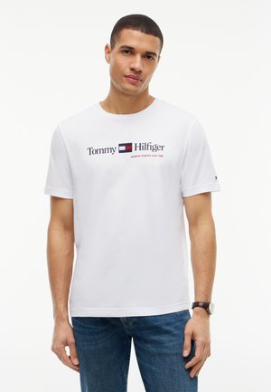 Homme portant un T-shirt blanc Tommy Hilfiger avec le texte du logo et un jean bleu, debout devant un fond blanc.